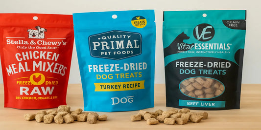 Three bags of freeze-dried dog treats in red, blue, and teal packaging are displayed on a wooden surface with treats scattered in front.